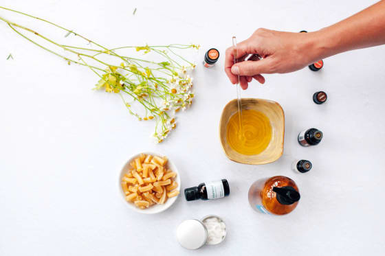 Overhead view of a hand stirring golden carrier oil in a wooden bowl, surrounded by chamomile flowers, wax pastilles, and amber essential oil bottles