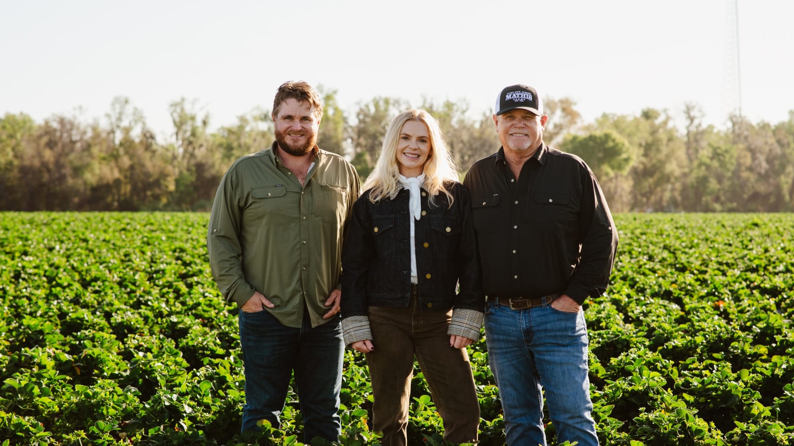Three smiling family members standing together in a lush green strawberry field on a farm.