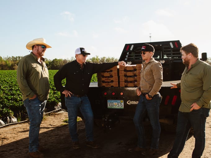 Four farmers in hats and jeans talk beside a flatbed truck loaded with produce boxes at a field's edge.