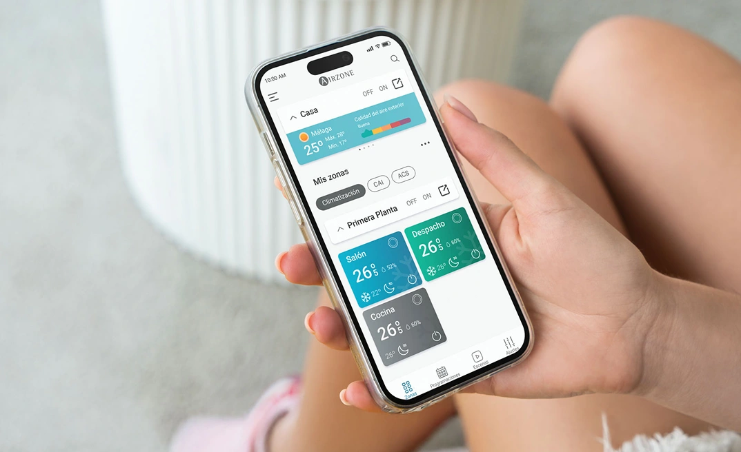 Persona sosteniendo un smartphone con la aplicación Airzone Cloud abierta mostrando la pantalla de inicio. Persona sosteniendo un smartphone con la aplicación Airzone Cloud abierta mostrando la pantalla de inicio.