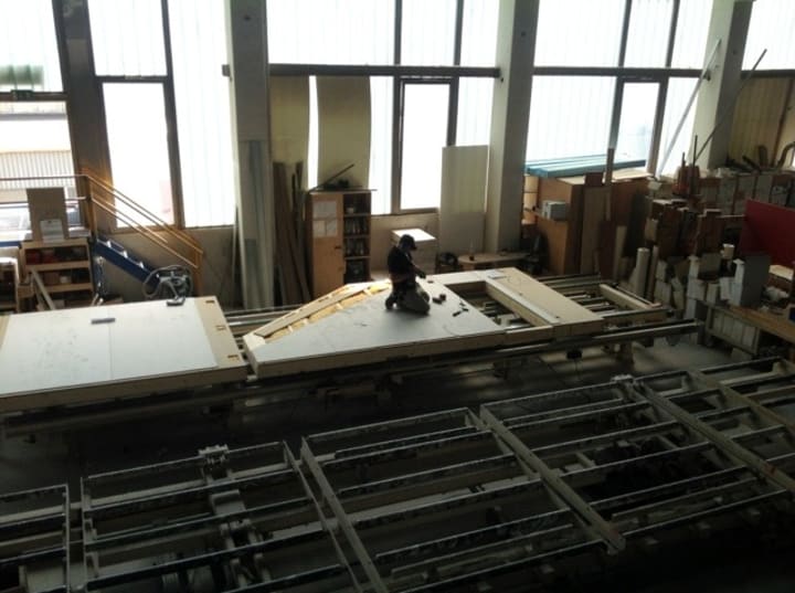 A man is kneeling on a wall piece with an electric screwdriver in his hand. The wall segment is itself on a conveyor belt.