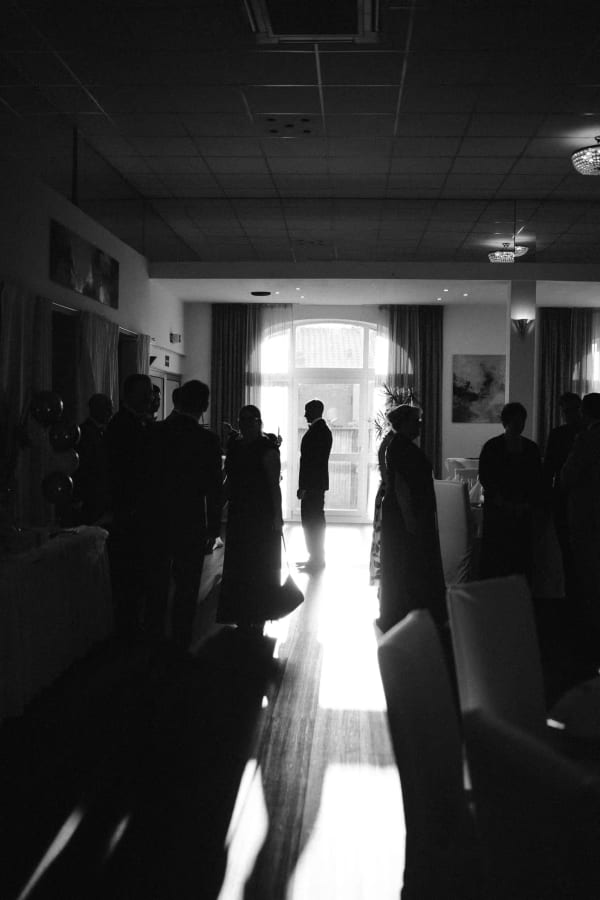 Emotionale Schwarz-Weiß-Fotografie von Hochzeitsgästen und dem Bräutigam als Silhouette im Gegenlicht einer großen Fenstertür während der Hochzeitsfeier im Hotel Hennies Isernhagen.