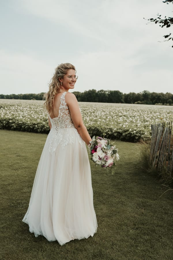 Eine lächelnde Braut mit Brille und einem Hochzeitskleid mit Spitzenrücken schaut über ihre Schulter zurück. Sie hält einen Brautstrauß mit weißen und lila Blüten. Sie steht auf der Wiese am Hotel Hennies Isernhagen in Hannover, mit einem großen blühenden Feld und einer Baumgrenze im Hintergrund. Ein rustikaler Zaun ist zu sehen.