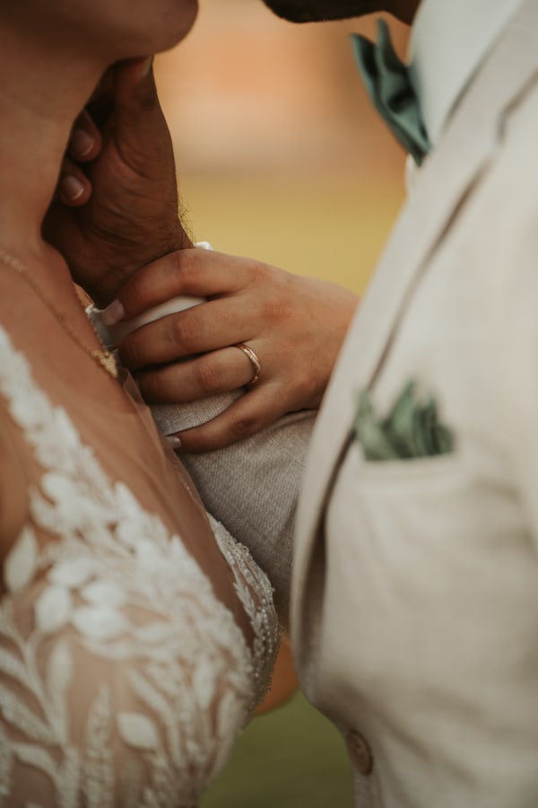 Detailaufnahme einer Hand mit Ehering, die zärtlich den Bräutigam berührt. Close-up von einem Hochzeitskleid mit floraler Spitze und einem hellen Hochzeitsanzug beim Hotel Hennies Isernhagen.