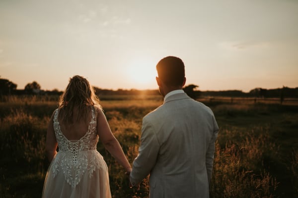 Rückenansicht von der Braut im verzierten Hochzeitskleid mit Spitzenrücken und Bräutigam, die Hand in Hand bei Sonnenuntergang über ein Feld in Isernhagen bei Hannover gehen.