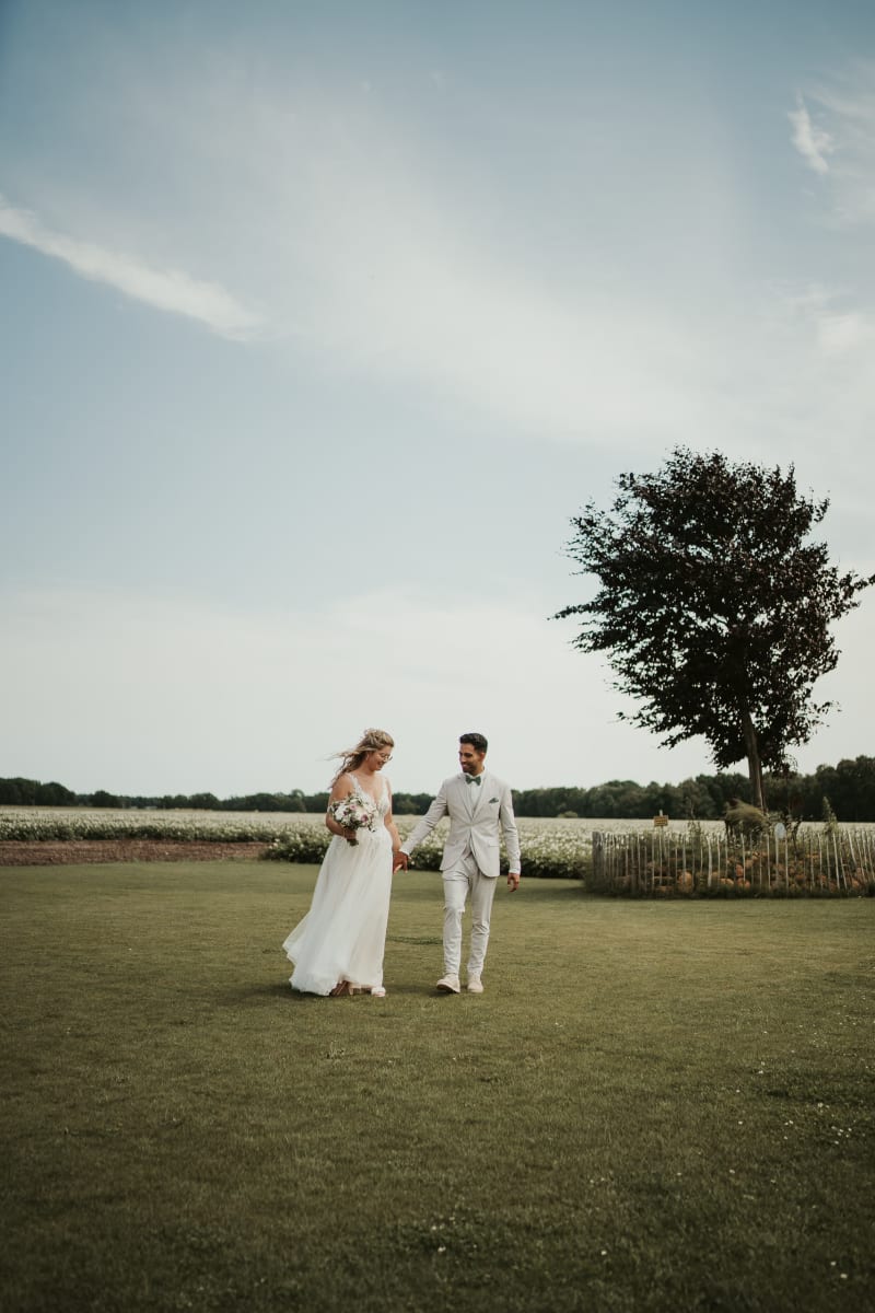 Weitwinkel-Hochzeitsfoto des Brautpaares beim Spaziergang über die große Rasenfläche mit einem markanten Baum im Hintergrund im Garten des Hotel Hennies Isernhagen.