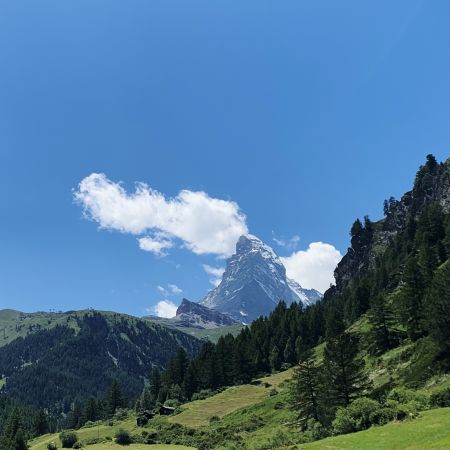 The Matterhorn as seen from Zermatt. In the foreground, green pastures and mountains. In the background, the Matterhorn stands out against a blue sky with a few clouds caressing its summit.