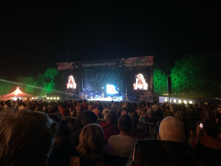 Angèle en concert sur la grande scène. on voit toute la grande scène. Sur chaque côté des écrans géants montre Angèle en train de chanter au micro. 40 000 personnes la regarde