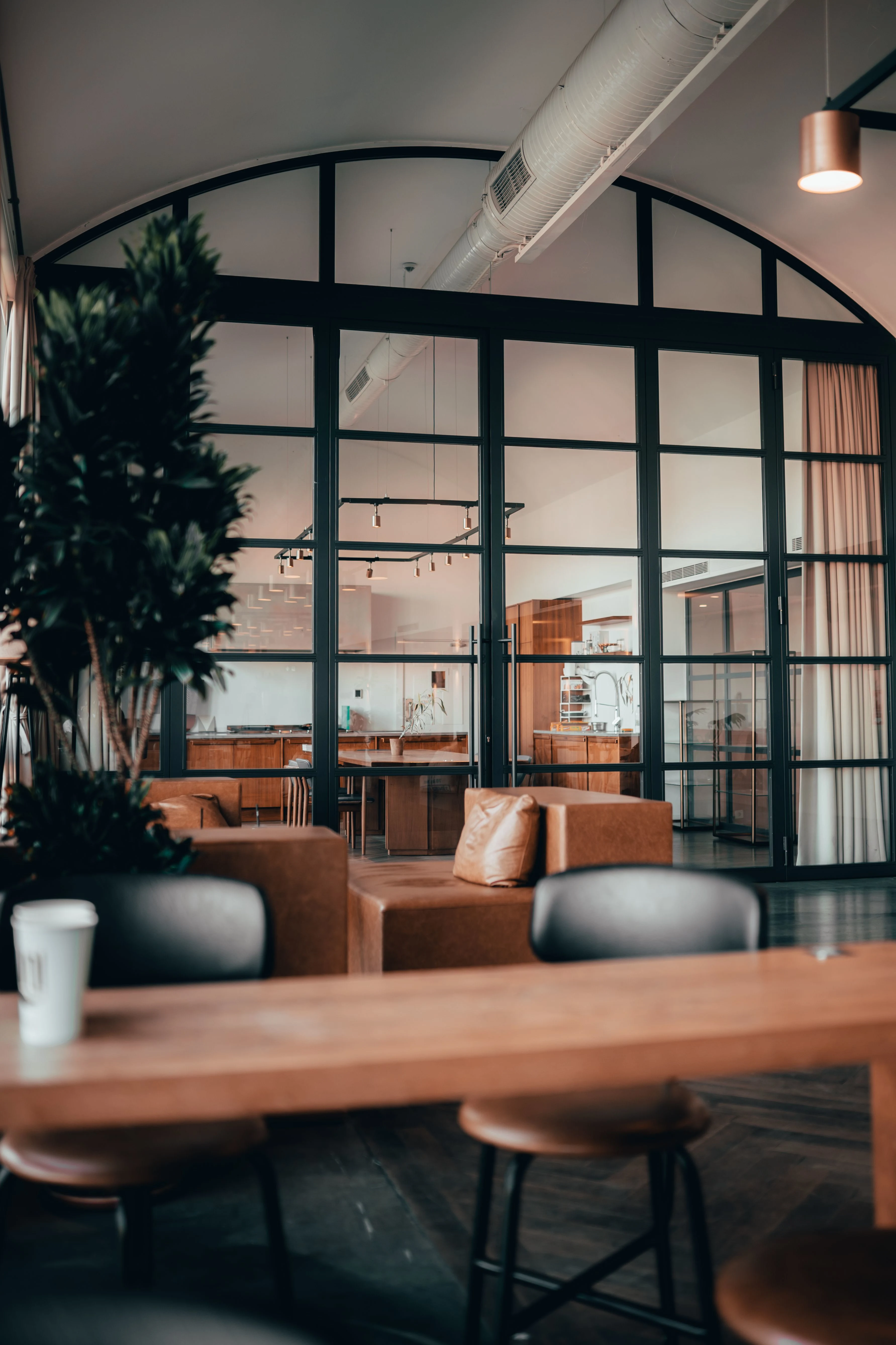 A coworking lounge with a wooden bar table and leather stools in front of arched black steel-framed glass walls