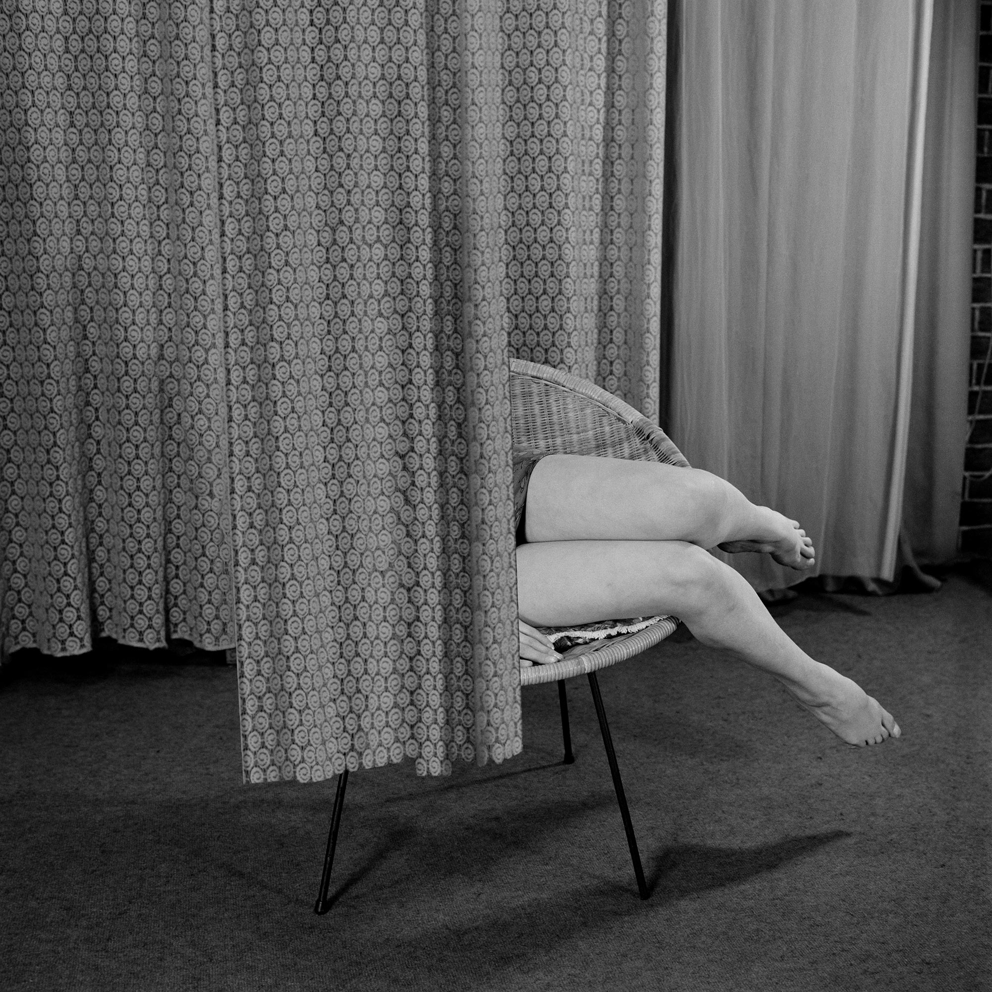 Black-and-white interior in which an extended patterned curtain conceals most of a seated woman, leaving her legs visible.