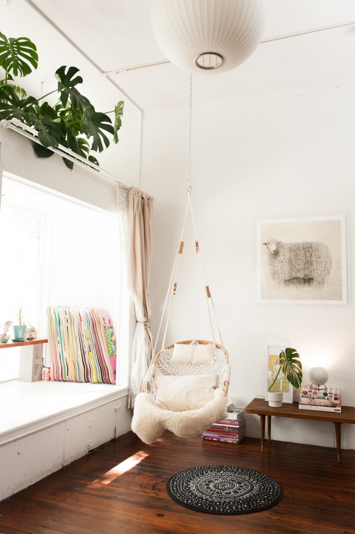 A Cobble Mountain hammock chair hanging in a room next to a large window letting in lots of sunlight. The hammock chair has a sheep skin blanket draped on it.