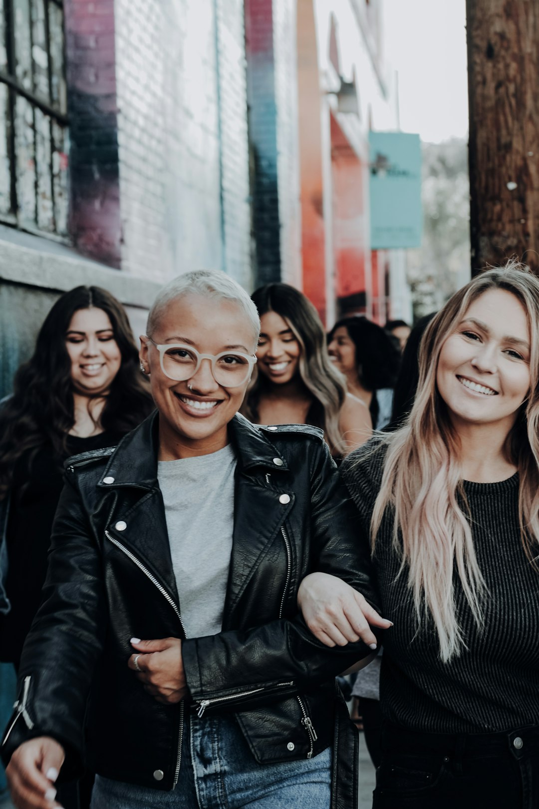 A diverse group of women walking together on a city street; the two women in the front have interlocking arms. 