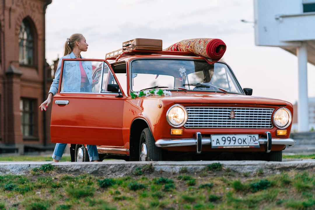 A woman in a denim jacket and long blonde hair in a ponytail is standing next to an orange car with the door open. It is in a parking lot in the daytime. She is looking at the items strapped to the top - a suitcase and a rolled up carpet. In the background are an old red building to the left and a white modern building to the right. 