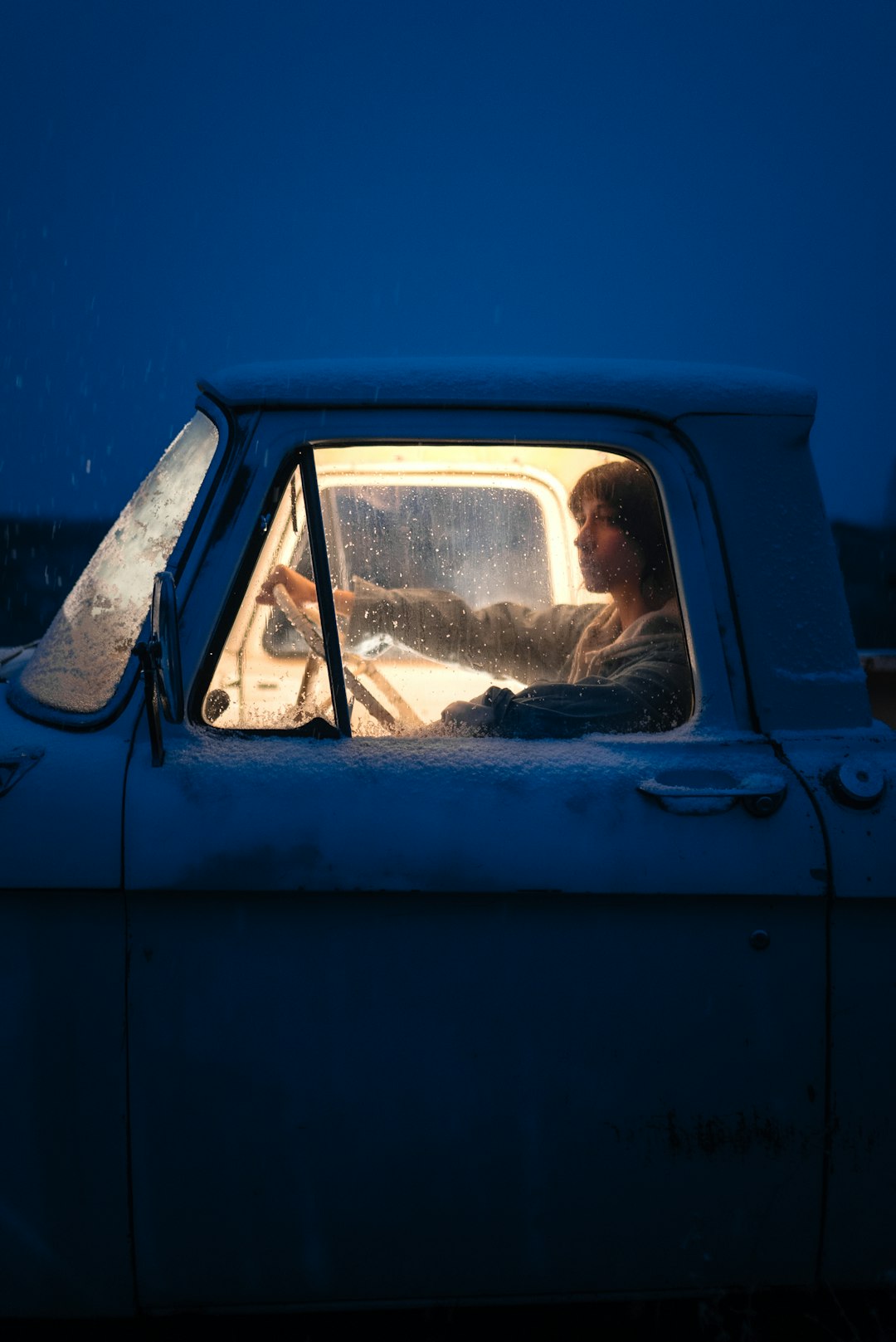 Teenager sitting in a truck at night with it's inside lights on looking forward with a hand on the wheel.