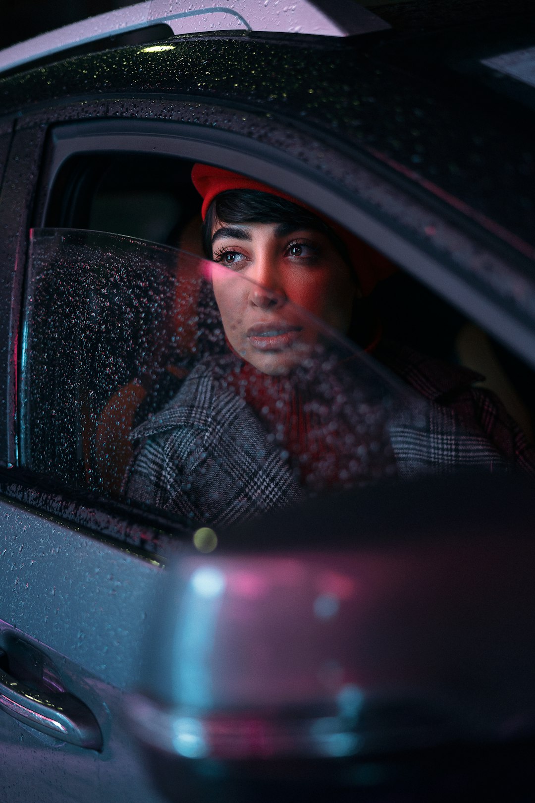 A woman in a car at night in the rain with her window partially rolled down. 