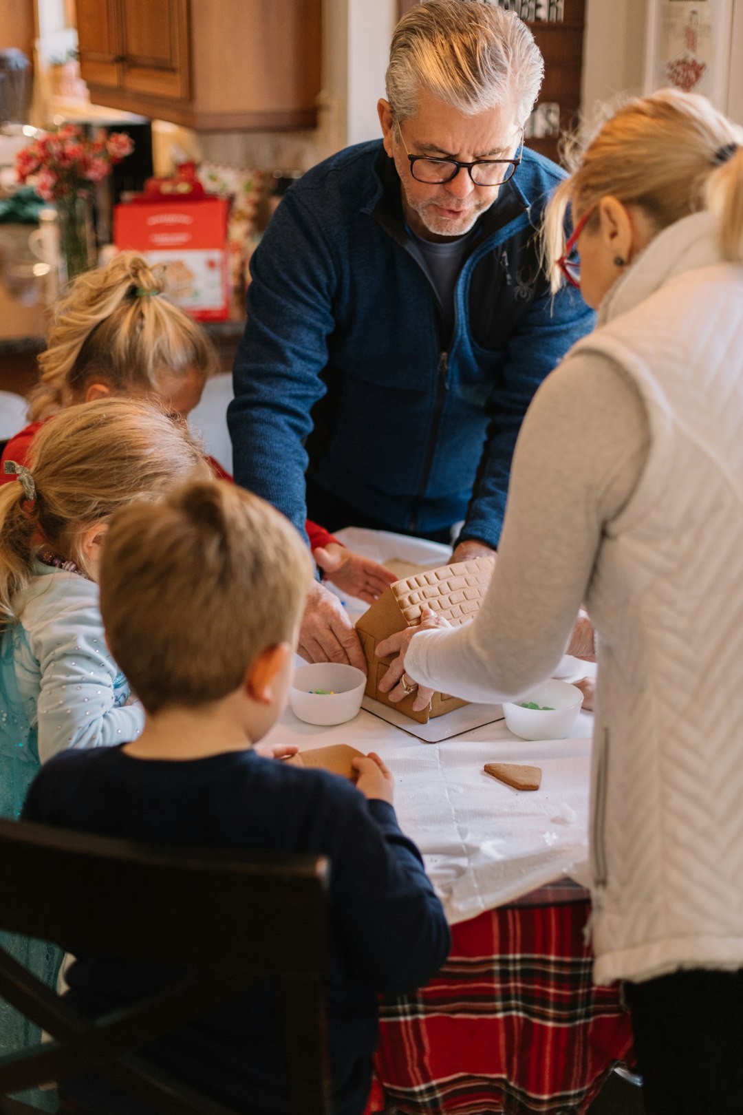 Grandparents are putting together a gingerbread house with their two young grandchildren, a boy and a girl both under 10 years old.