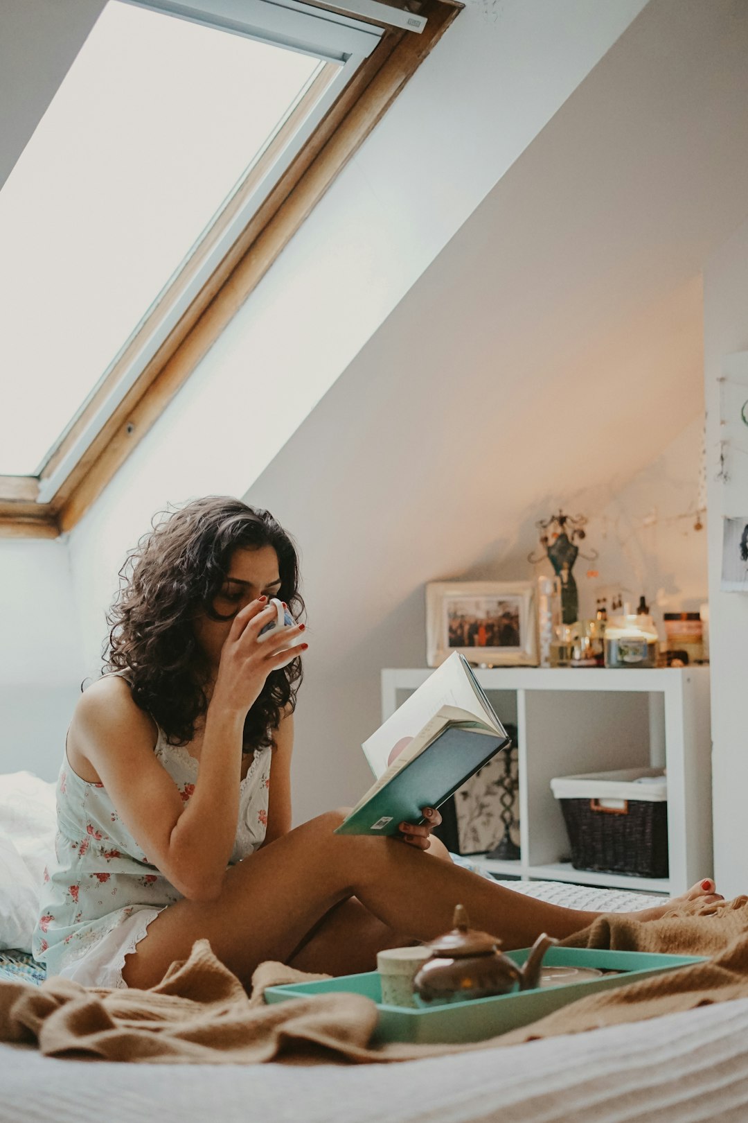 Woman in sleep clothes sitting on the bed sipping a cup of tea while reading a book