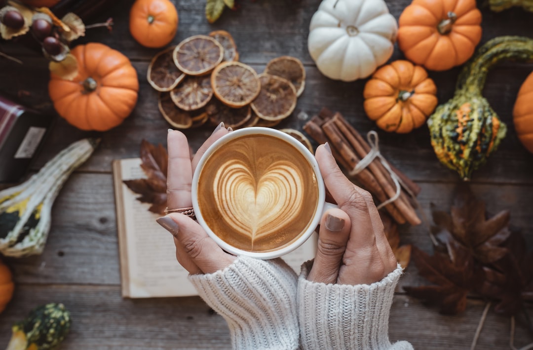 woman holding a cappuccino with a heart in it. Background has pumpkins, sliced oranges, and other fall objects.