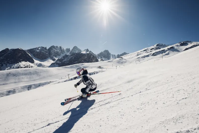 Eine Person fährt Ski den Hang hinunter, umgeben von schneebedeckten Bergen und klarem Himmel.