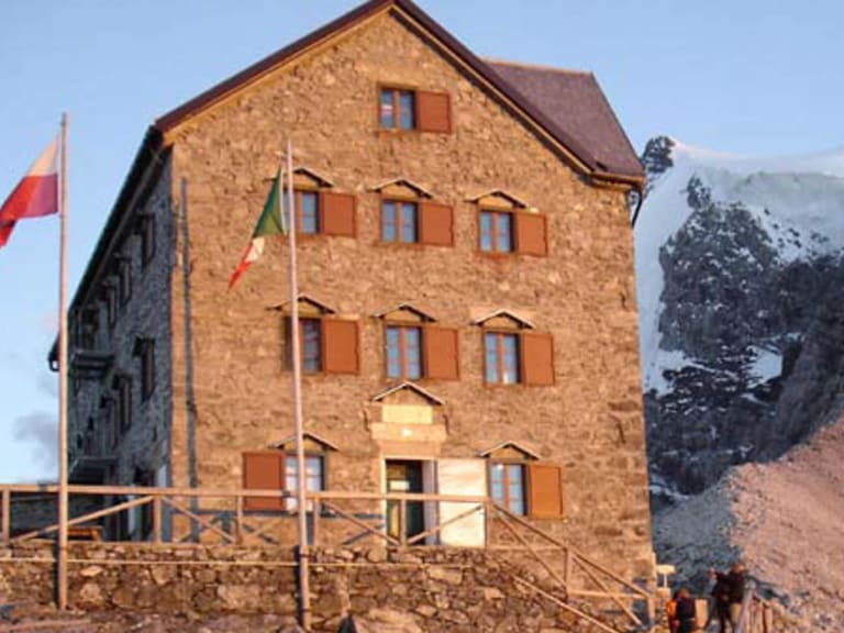 Bergwanderer vor der Julius Payer Hütte auf einem schmalen Felsgrat mit schneebedeckten Gipfeln im Hintergrund.