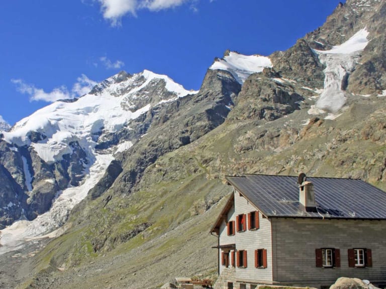 Die Chamanna da Boval steht auf steinigem Untergrund vor vergletscherten Gipfeln der Bernina-Alpen.