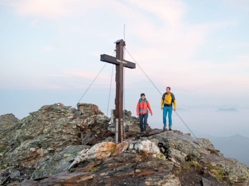 Zwei Bergwanderer stehen neben einem hölzernen Gipfelkreuz in den Sarntaler Alpen über einem Nebelmeer.
