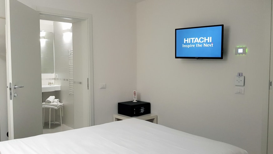 Modernes Doppelzimmer im Hotel Aurora Duino mit weißem Bett, Flachbildfernseher und Safe auf Beistelltisch.