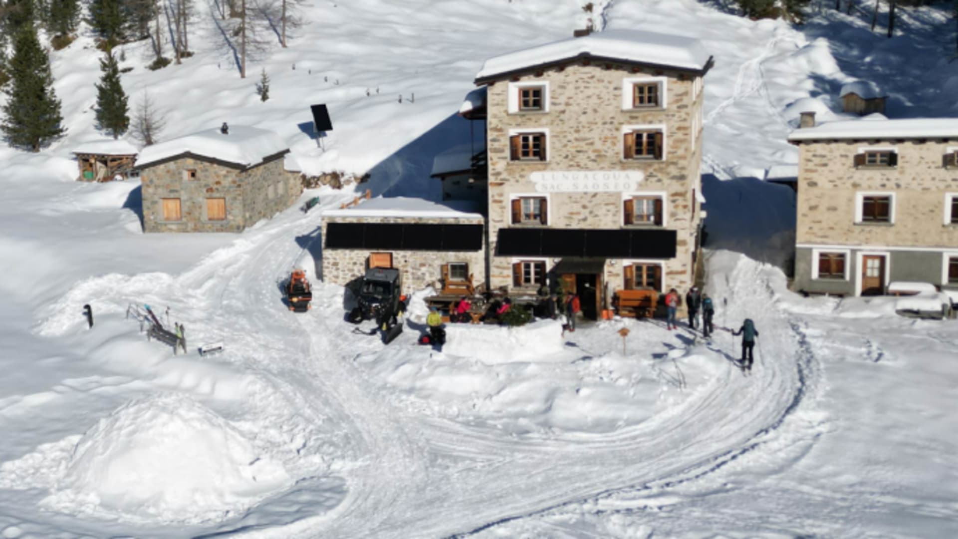 Luftaufnahme des Rifugio Saoseo im Val de la Camp – Skitourengeher im Aufstieg, verschneite Steingebäude und Nadelwald