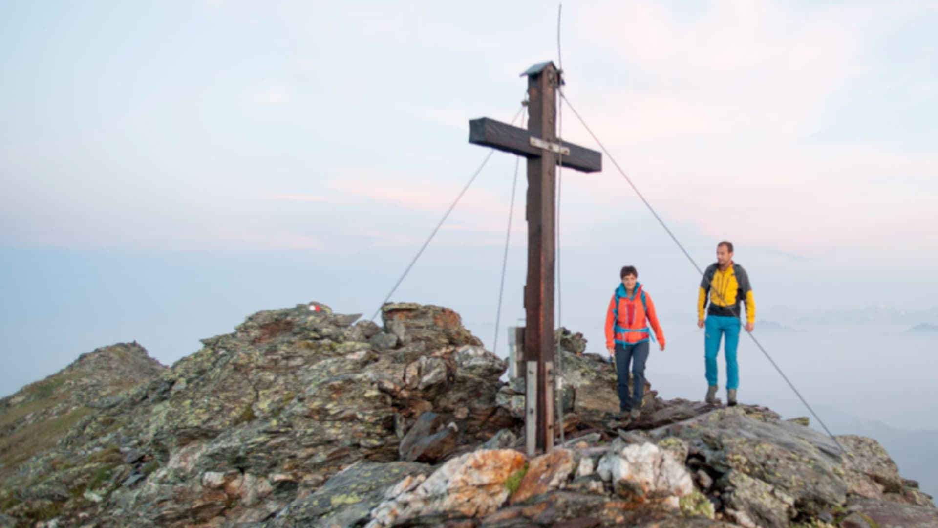 Zwei Bergwanderer stehen neben einem hölzernen Gipfelkreuz in den Sarntaler Alpen über einem Nebelmeer.