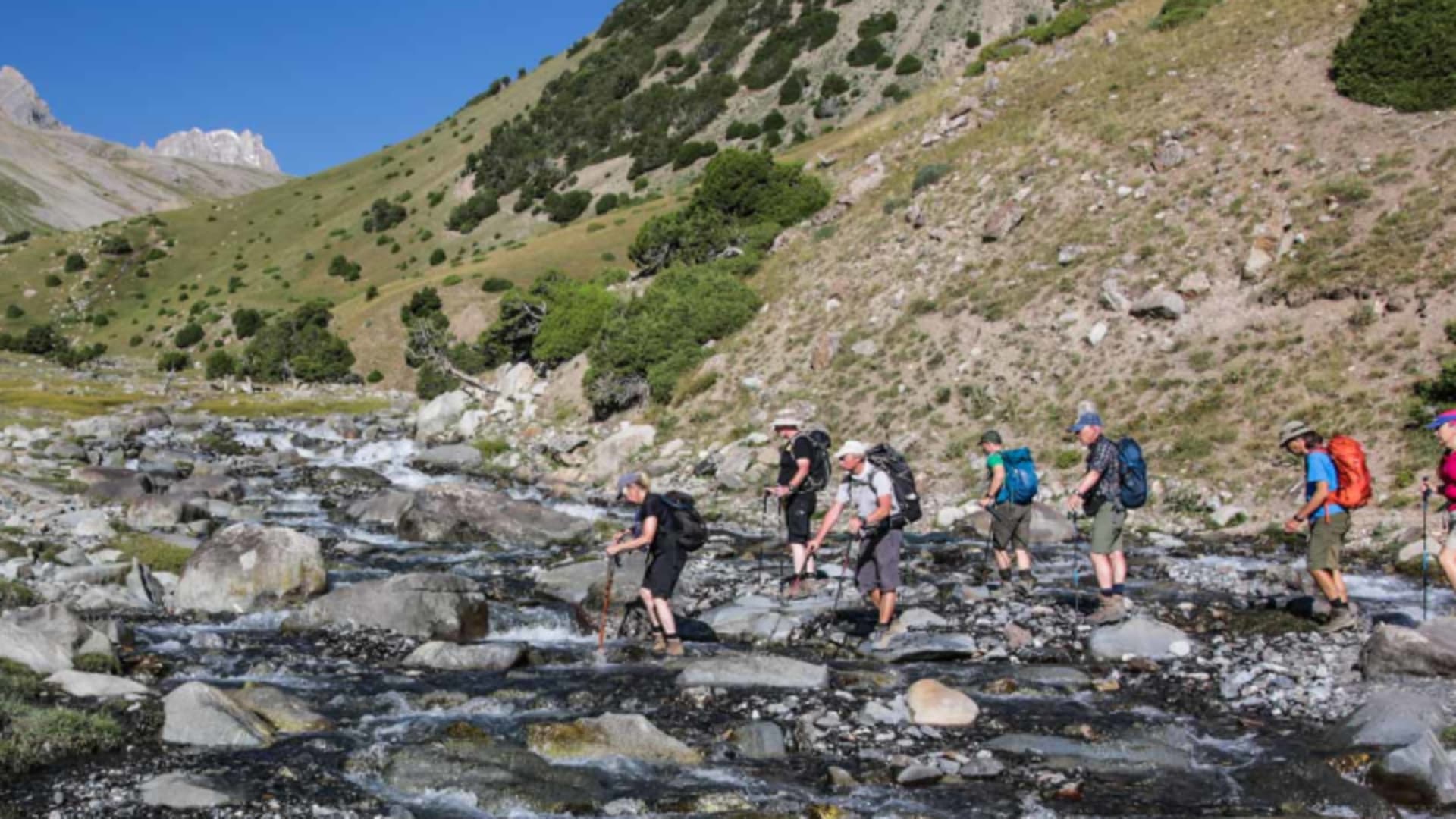 Trekkergruppe überquert einen flachen Gebirgsbach in Kirgistan mit Rucksäcken und Wanderstöcken auf Steinen.