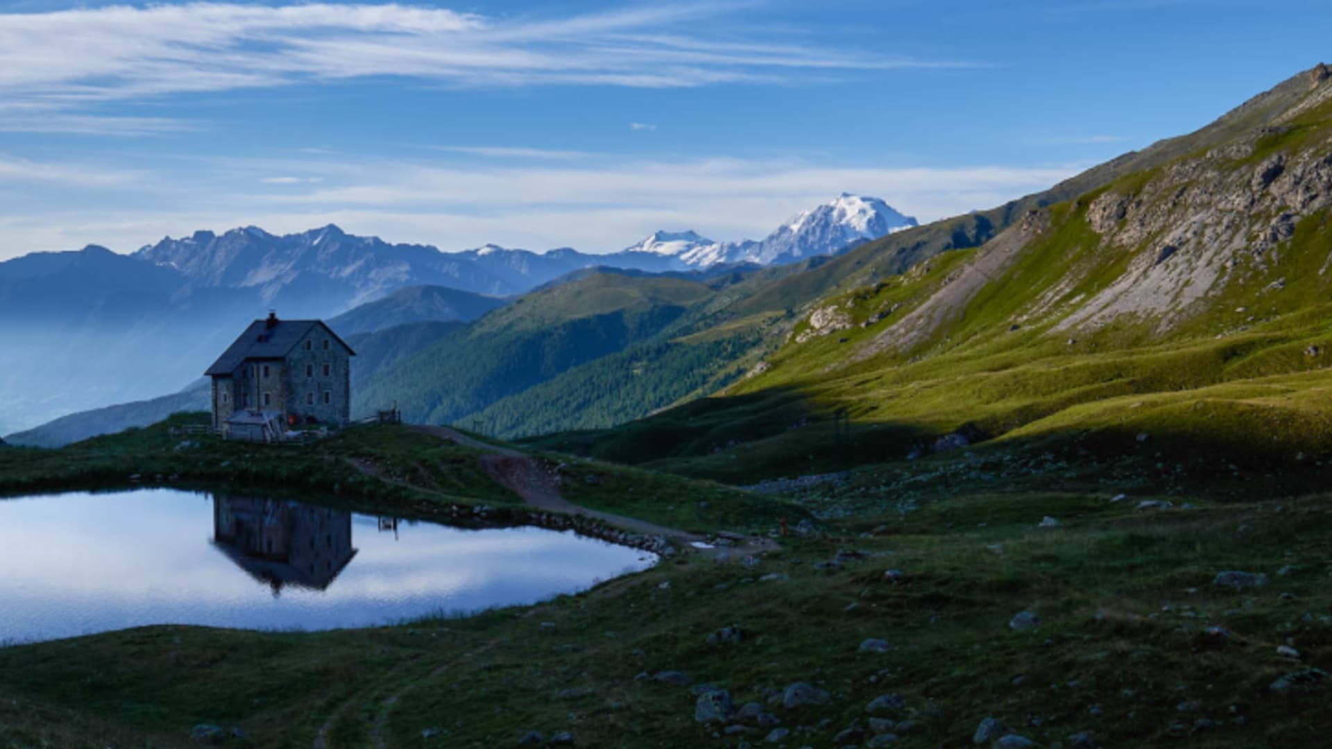 Berghütte im Engadin mit herzförmigem Bergsee im Vordergrund zur Blauen Stunde, schneebedeckte Gipfel im Hintergrund.