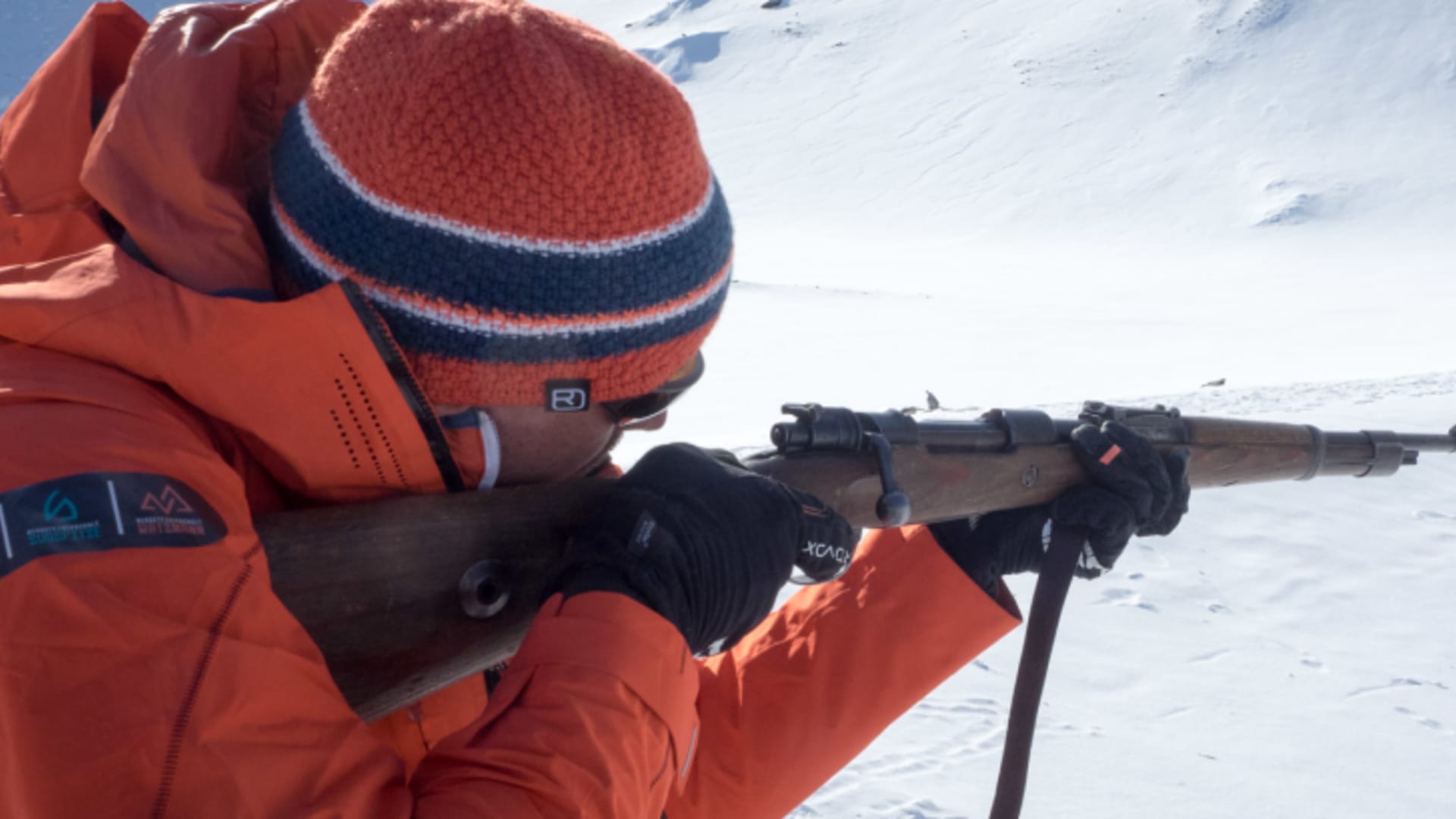 Skitourengeher in oranger Winterjacke zielt mit Repetiergewehr auf Schneehang in Spitzbergen.
