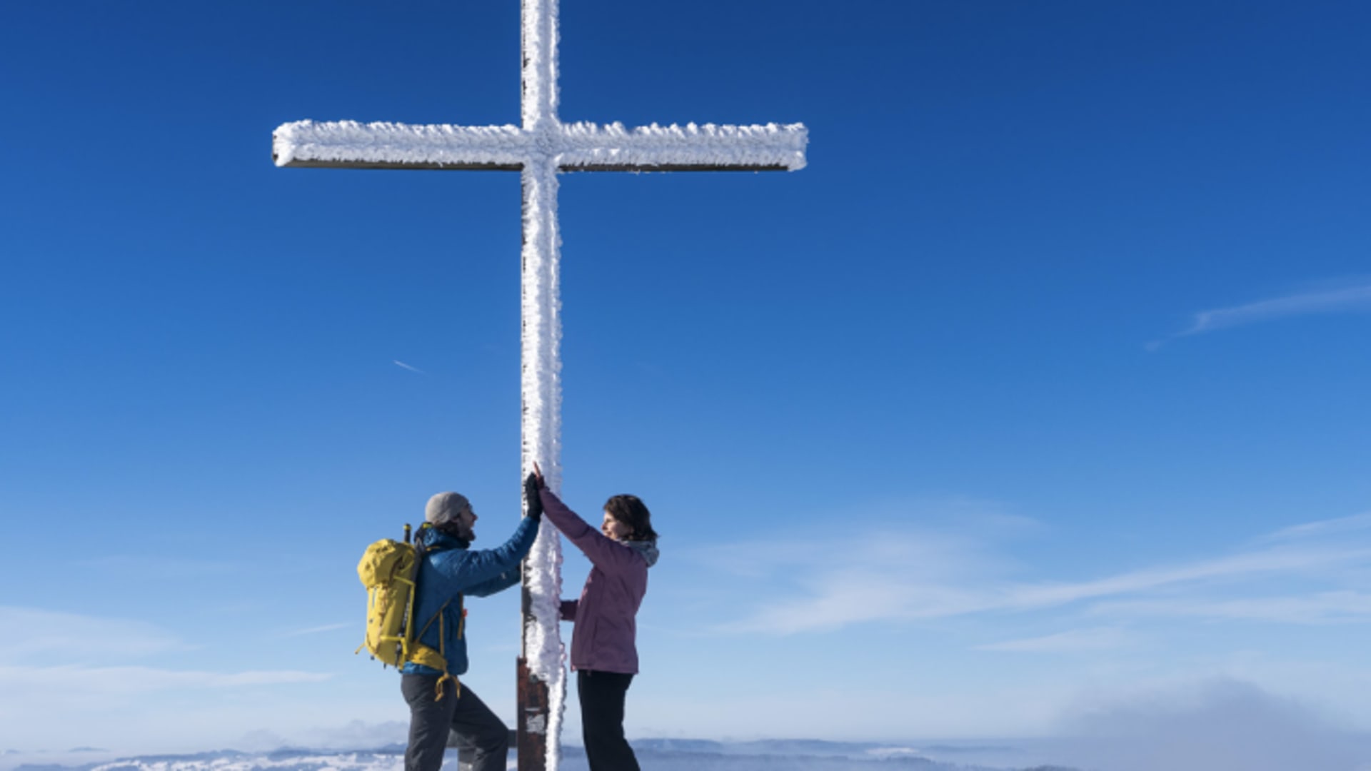 Zwei Winterwanderer berühren ein raureifbedecktes Metallgipfelkreuz bei Oberstaufen in den Allgäuer Alpen.