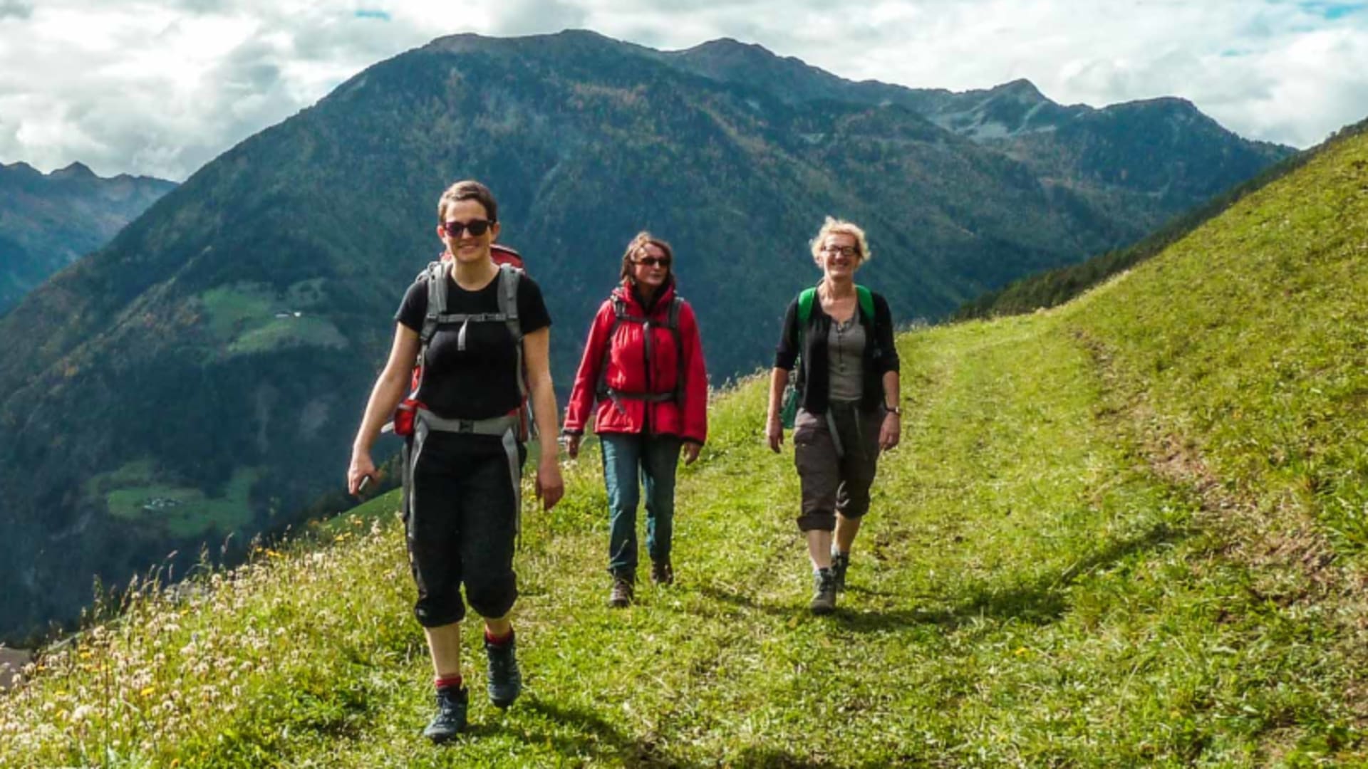 Drei Bergwanderer steigen mit Rucksäcken über eine grüne Almwiese bergauf, im Hintergrund bewaldete Berggipfel in Südtirol.
