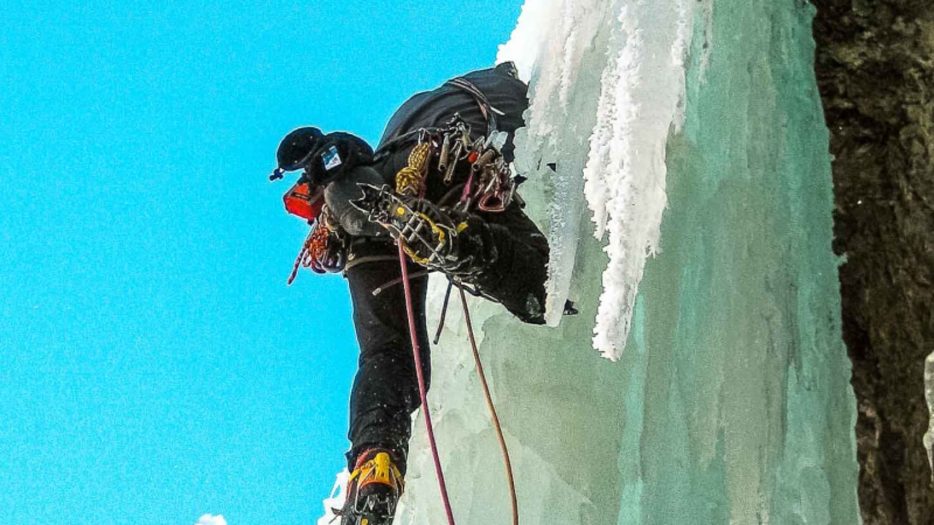 Eiskletterer mit Steigeisen und Eisgerät an senkrechter türkis-blauer Eiswand in den Alpen, begleitet durch eine Bergführer-Führung