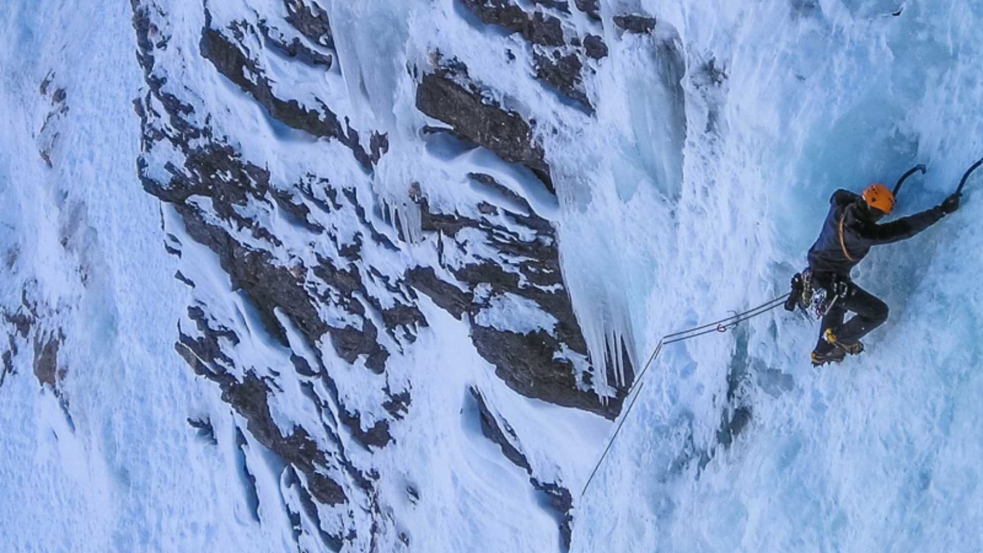 Eiskletterer mit orangem Helm und Eisäxten steigt senkrechte Eiswand in den Dolomiten empor, Seil führt zu Zwischensicherung