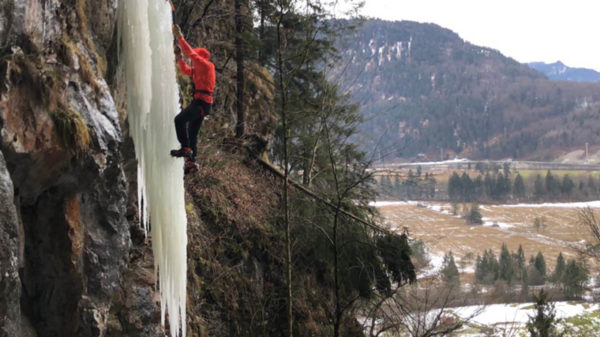 Eiskletterer in roter Jacke greift mit beiden Händen in vertikale Eissäule bei Oberau, Talblick.