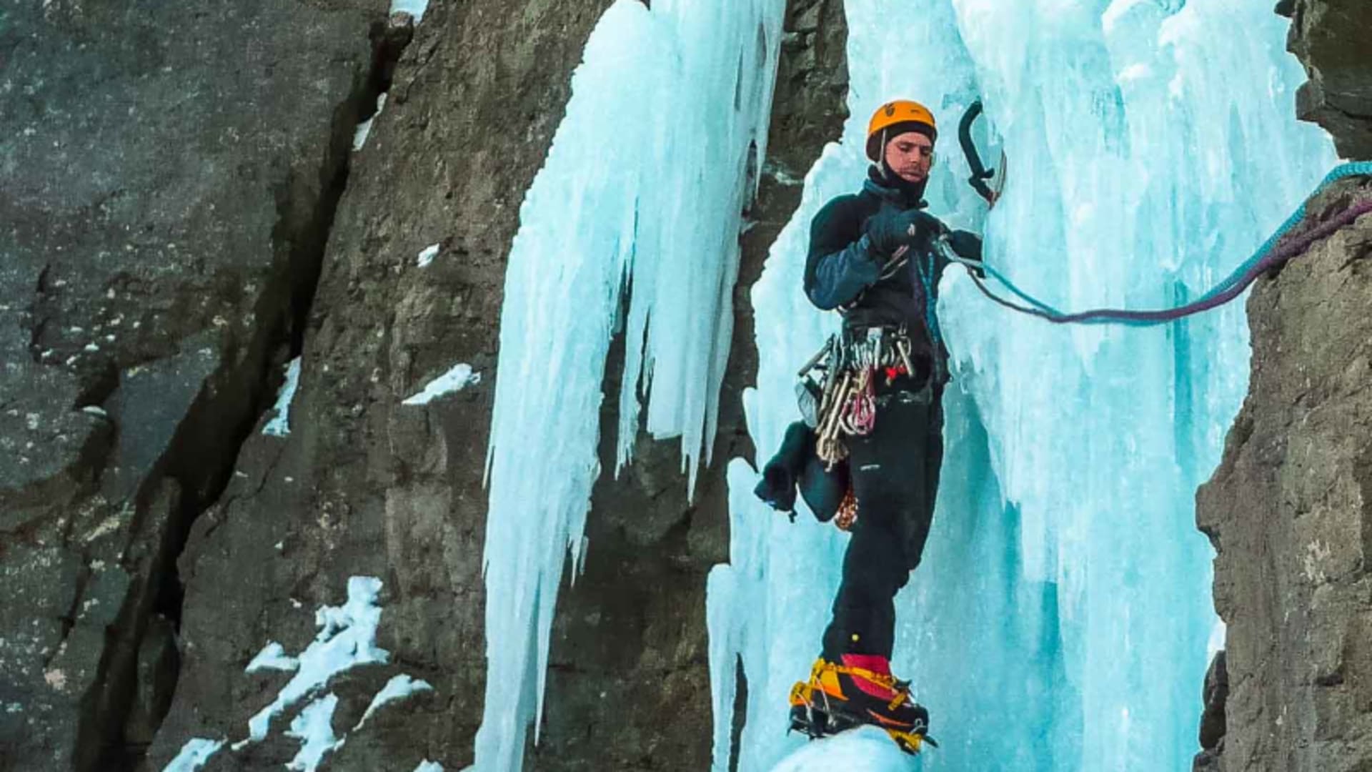 Eiskletterer steht mit Helm, Steigeisen und Eispickel vor blauem Eiswasserfall beim Eiskletterfestival in Kandersteg.
