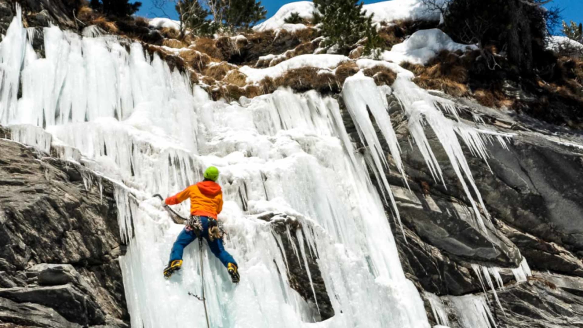 Eiskletterer mit Rücken zur Kamera steigt gefrorenen Wasserfall in der Schweiz mit Eisgeräten und Steigeisen empor.