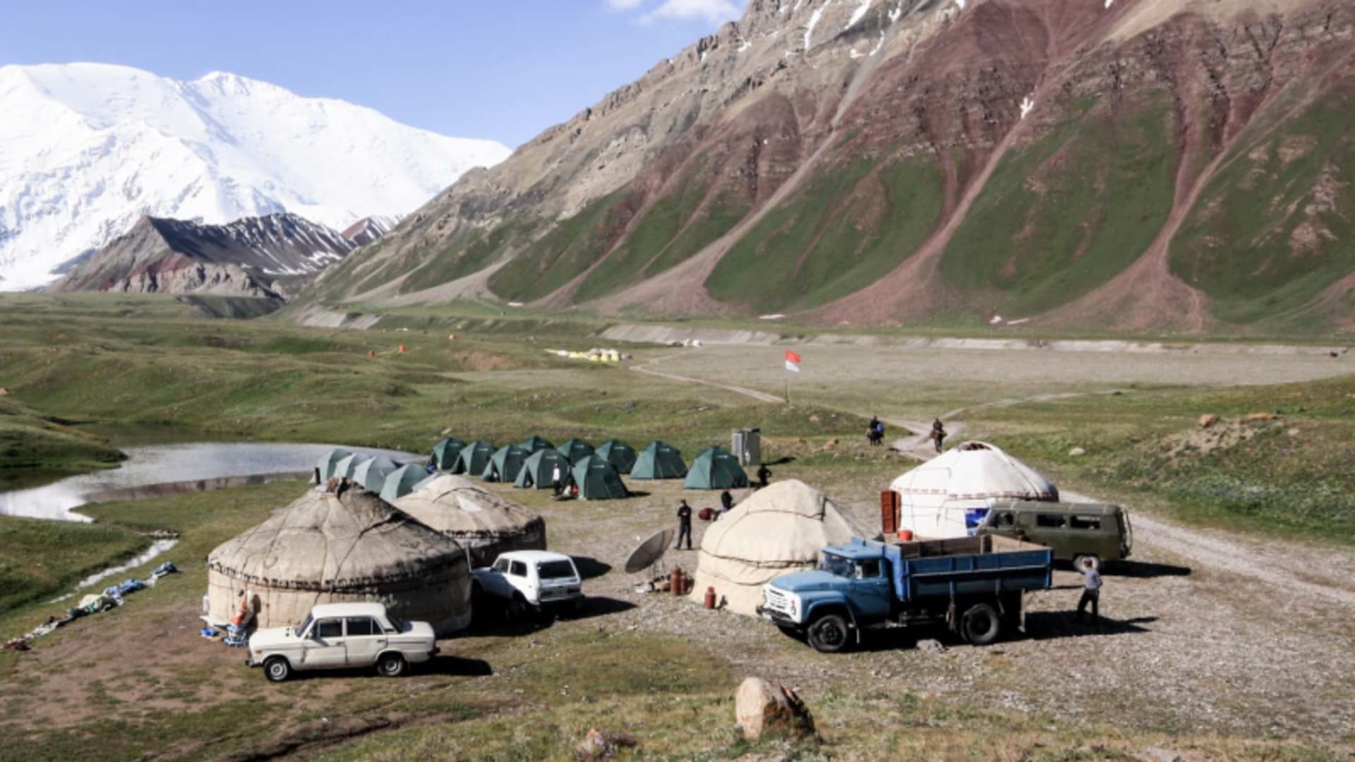 Expedition Pik Lenin: Basislager Achik Tasch mit Jurten, Zelten und Fahrzeugen im Hochtal vor verschneiten Gipfeln.