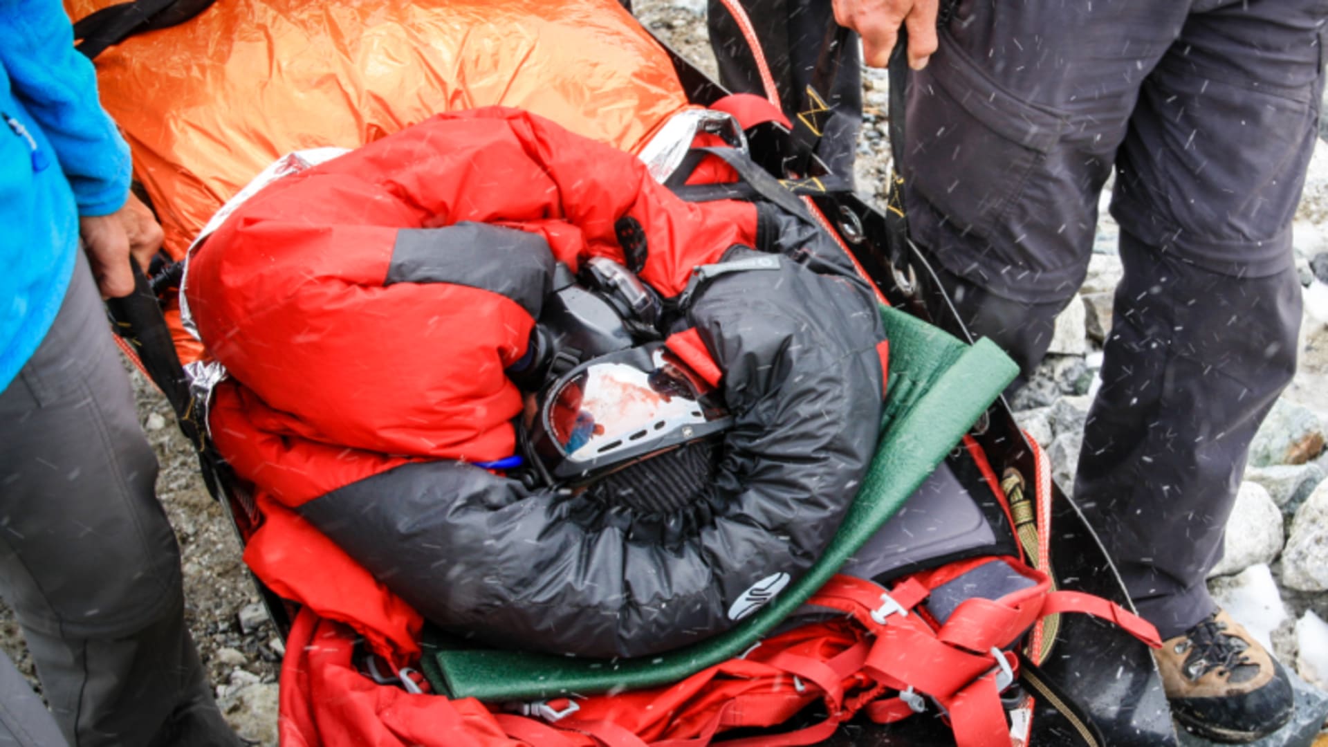 Expedition-Rettung am 8000er: Höhenbergsteiger auf roter Akia-Trage fixiert, zwei Helfer auf Felsblöcken bei Schneefall.