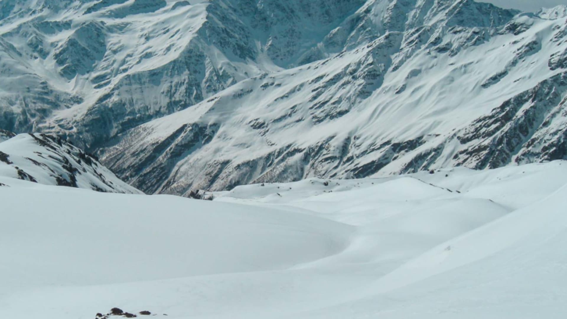 Expedition am Elbrus: ein Höhenbergsteiger mit Skiern auf weitem Schneefeld vor verschneiter Bergkette.