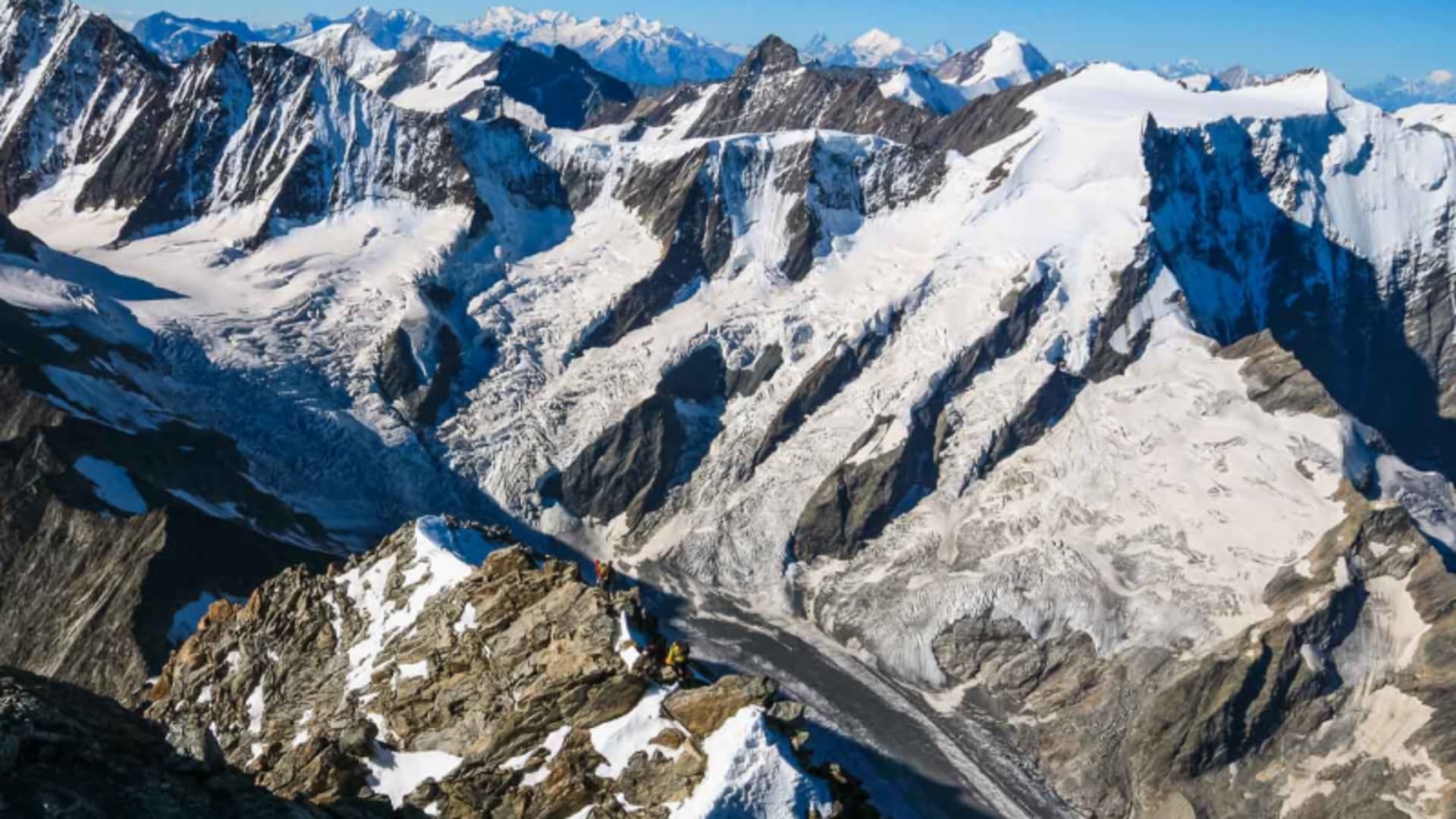 Hochtour im Berner Oberland: Bergsteigergruppe auf felsigem Schnee-Grat mit Gletscherbecken und Bergkette im Hintergrund.