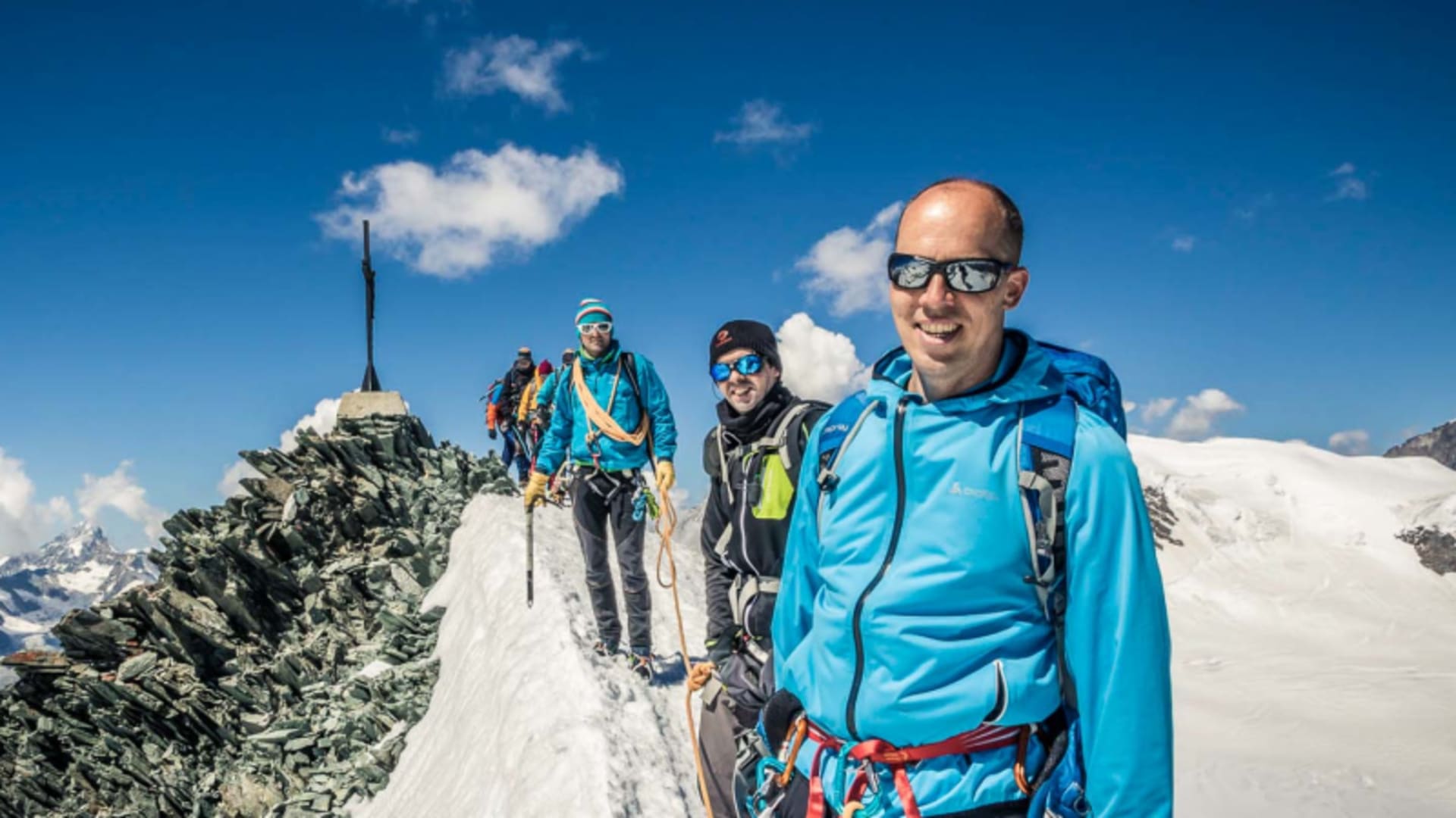 Drei Bergsteiger als Seilschaft auf dem Schneegrat des Allalinhorn mit Gipfelkreuz im Hintergrund.