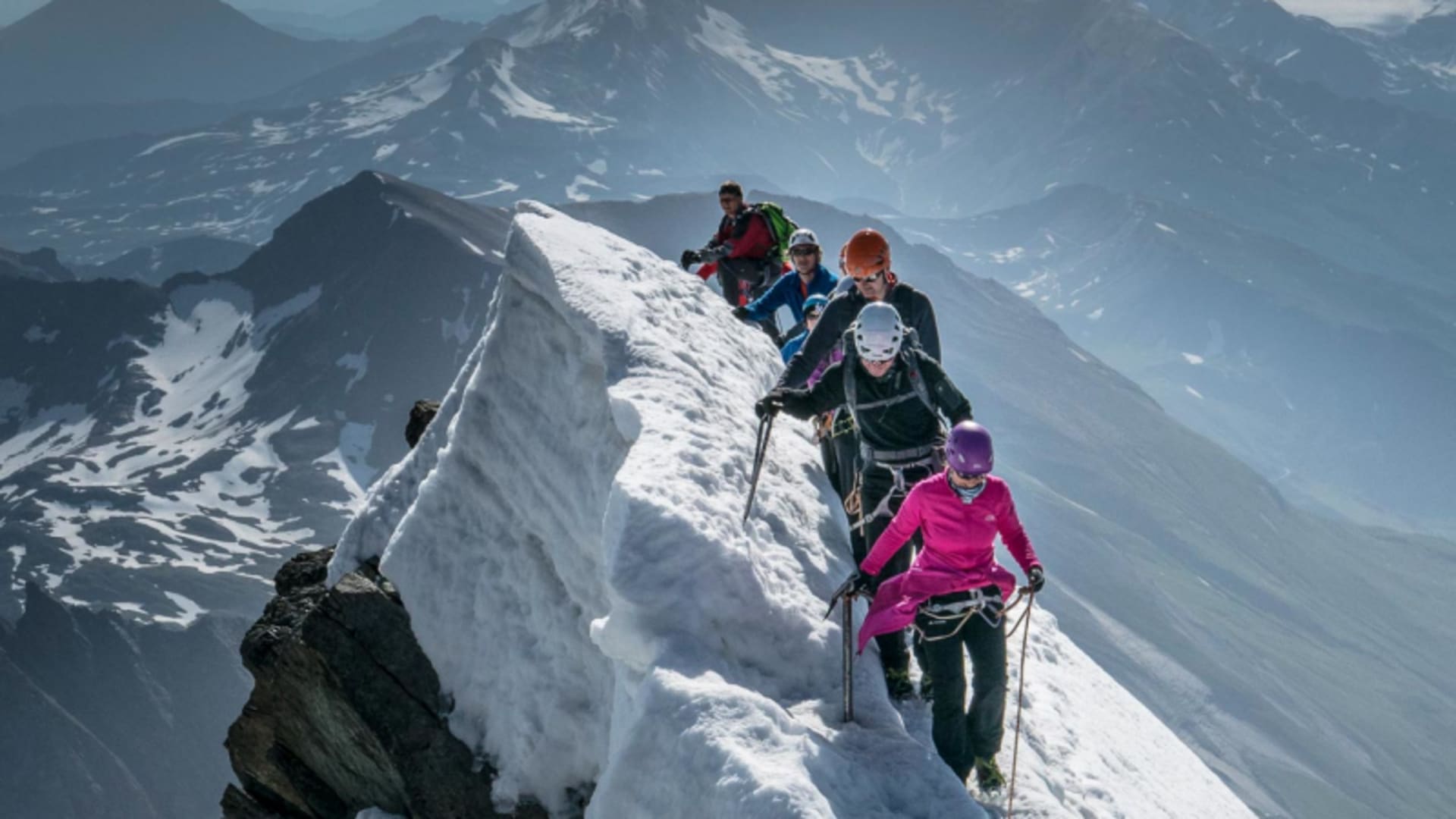 Fünf Bergsteiger traversieren am verschneiten Gipfelgrat des Großglockner mit Seil, Helmen und Klettergurten gesichert.
