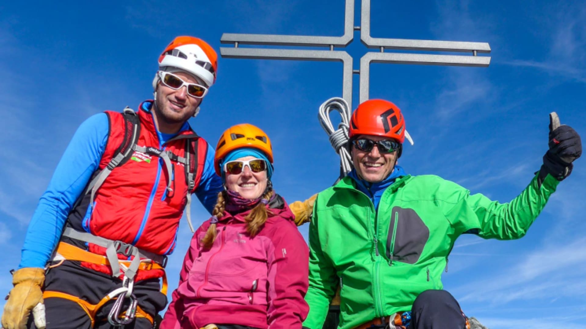 Drei Bergsteiger mit Helm und Klettergurt posieren am Gipfelkreuz beim Nadelgrat im Wallis bei blauem Himmel.