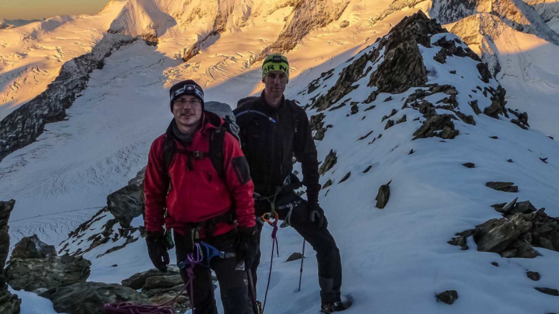 Hochtour im Berner Oberland: Zwei Bergsteiger mit Klettergurt und Eisgerät pausieren auf einem schmalen Schneegrat bei Morgenrot.