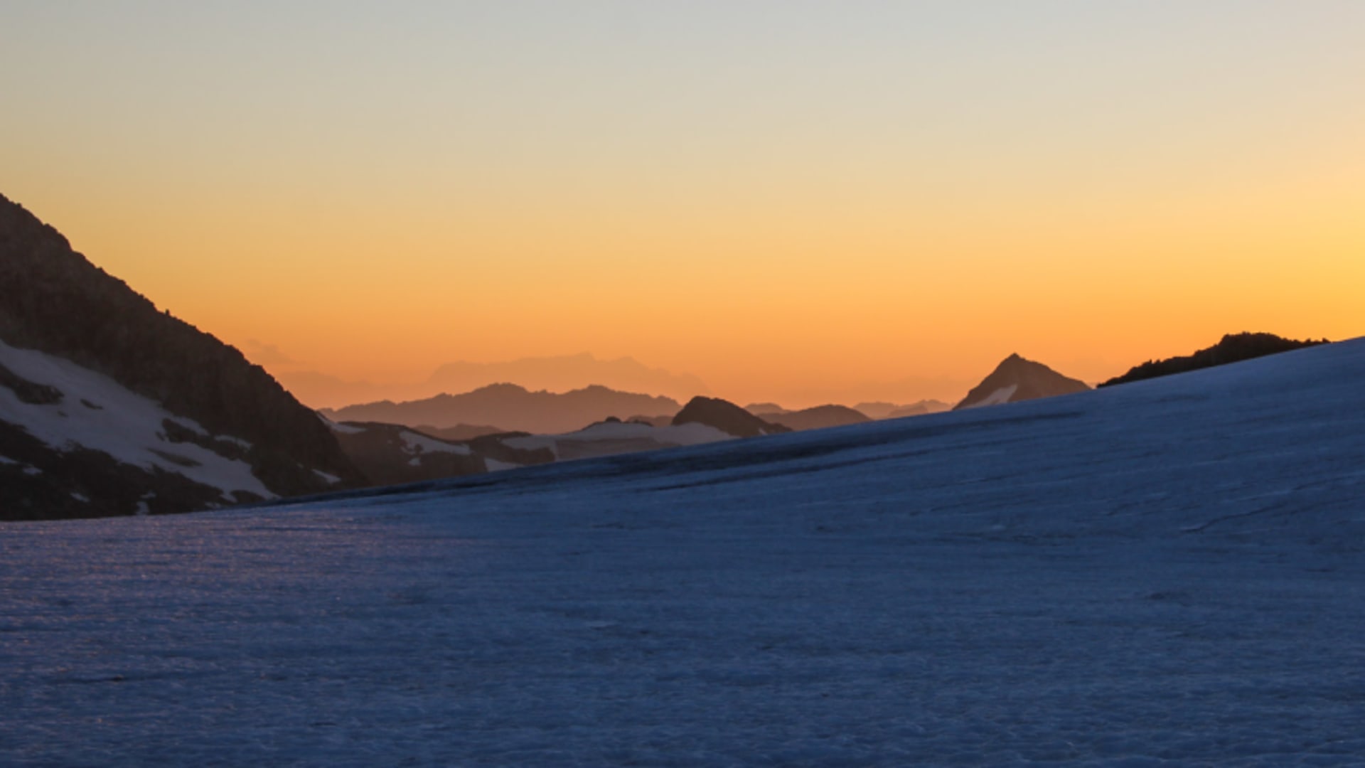Schneebedeckter Hang am Großvenediger bei Sonnenaufgang mit orangegelbem Lichtschein und Bergsilhouetten im Hintergrund.