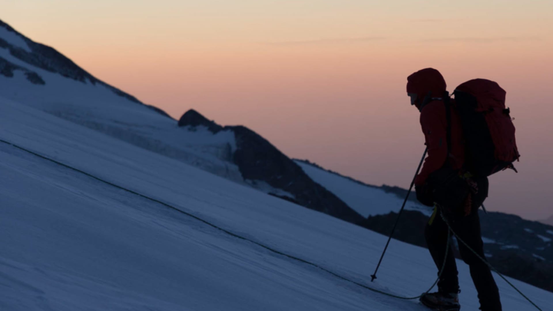 Bergsteiger steigt mit Steigeisen und Pickel am Seil einen steilen Schneehang bei Sonnenaufgang in den Walliser Alpen auf.