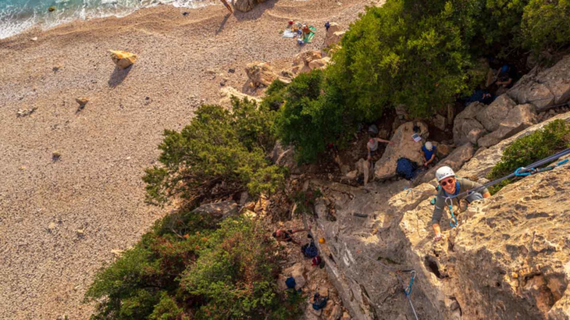 Kletterer mit weißem Helm steigt an Kalkfelswand direkt über türkisblauer Meeresbucht auf – Felsklettern in Sardinien mit Gruppe